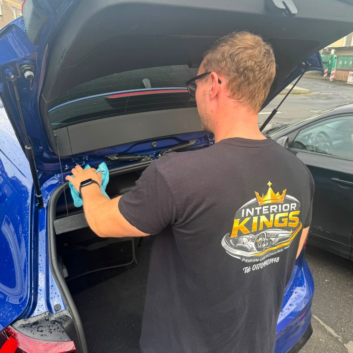 Aufbereiter von InteriorKings im Firmenshirt reinigt den Kofferraum eines blauen VW Golf R von Hand